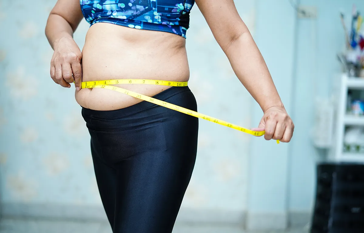 Neprepoznatljiva žene srednje do starije životne dobi, obučena u sportsku odjeću, metrom mjeri opseg masnog tkiva na području trbuha; koncept prehrana u menopauzi