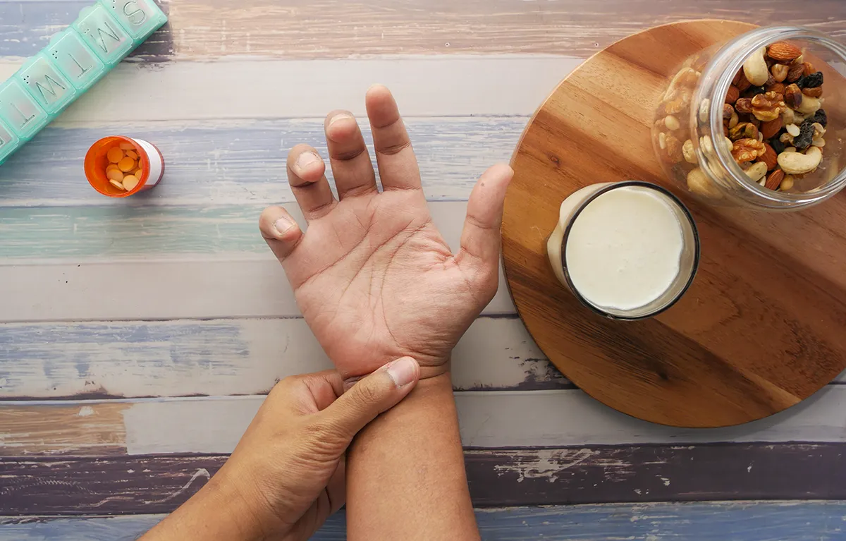 reumatoidni-artritis-prehrana Muškarac s bolnim rukama iznad stola s orašastim plodovima, mliječnim napitkom i kutijom tableta; koncept prehrana reumatoidni artritis