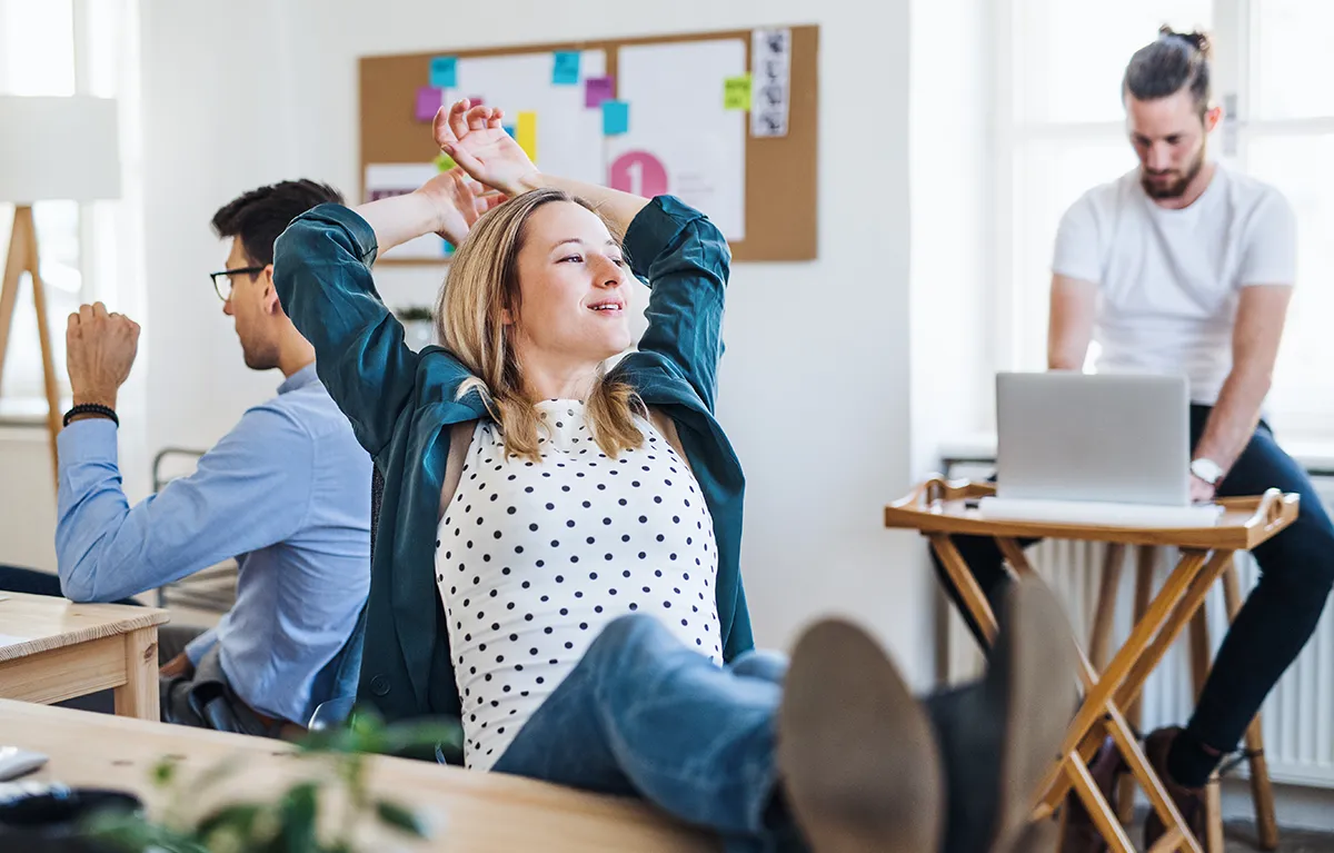 Mlađi zaposlenici u uredu, zaposlenica odmara s nogama na stolu i ispruženih ruku; koncept CVS sindrom