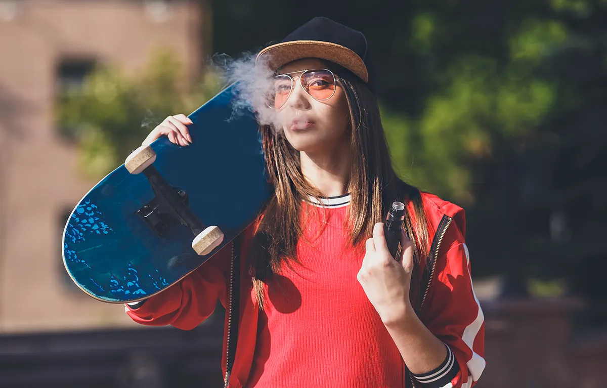 Djevojka gleda u kameru, drži skateboard i koristi električnu cigaretu; koncept žuti zubi kao posljedica duhana