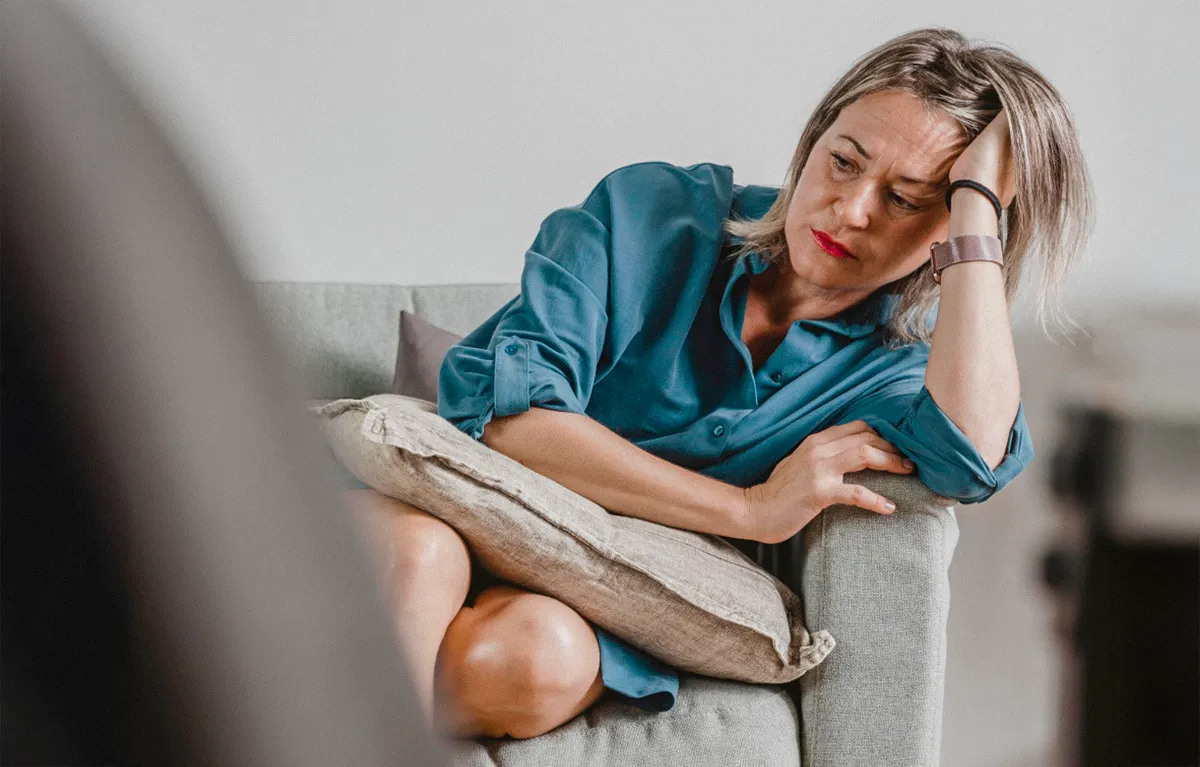 Umorna i tužna žena sjedi na kauču skupljenih nogu i glave naslonjene na ruku; koncept vježbanje u perimenopauzi