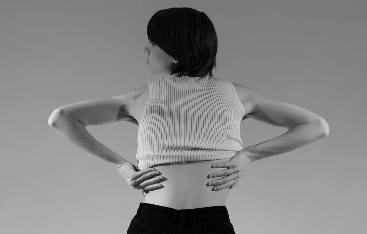 Žena u bijeloj potkošulji rukama se drži za bolni donji dio leđa; koncept lijek protiv fibromialgije