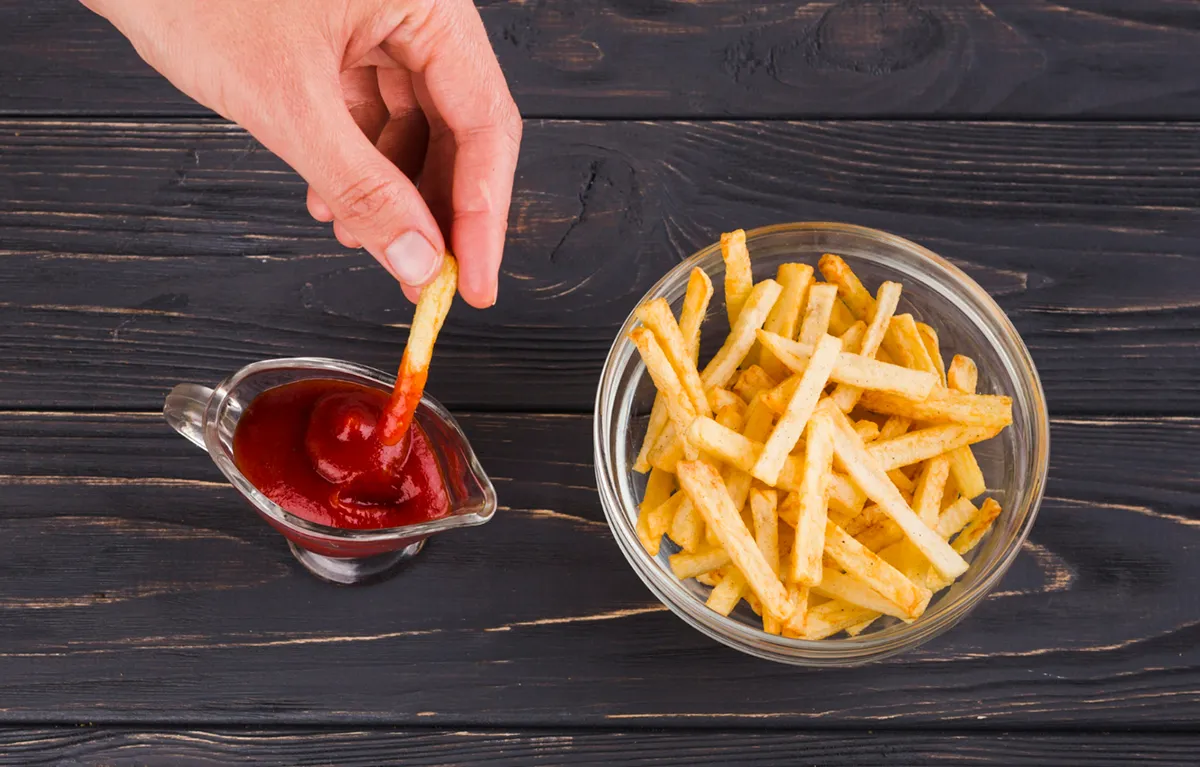 Pomfrit u staklenoj zdjelici i ruka koja krumpirić stavlja u kečap; koncept pomfrit i dijabetes tipa 2, koja je povezanost