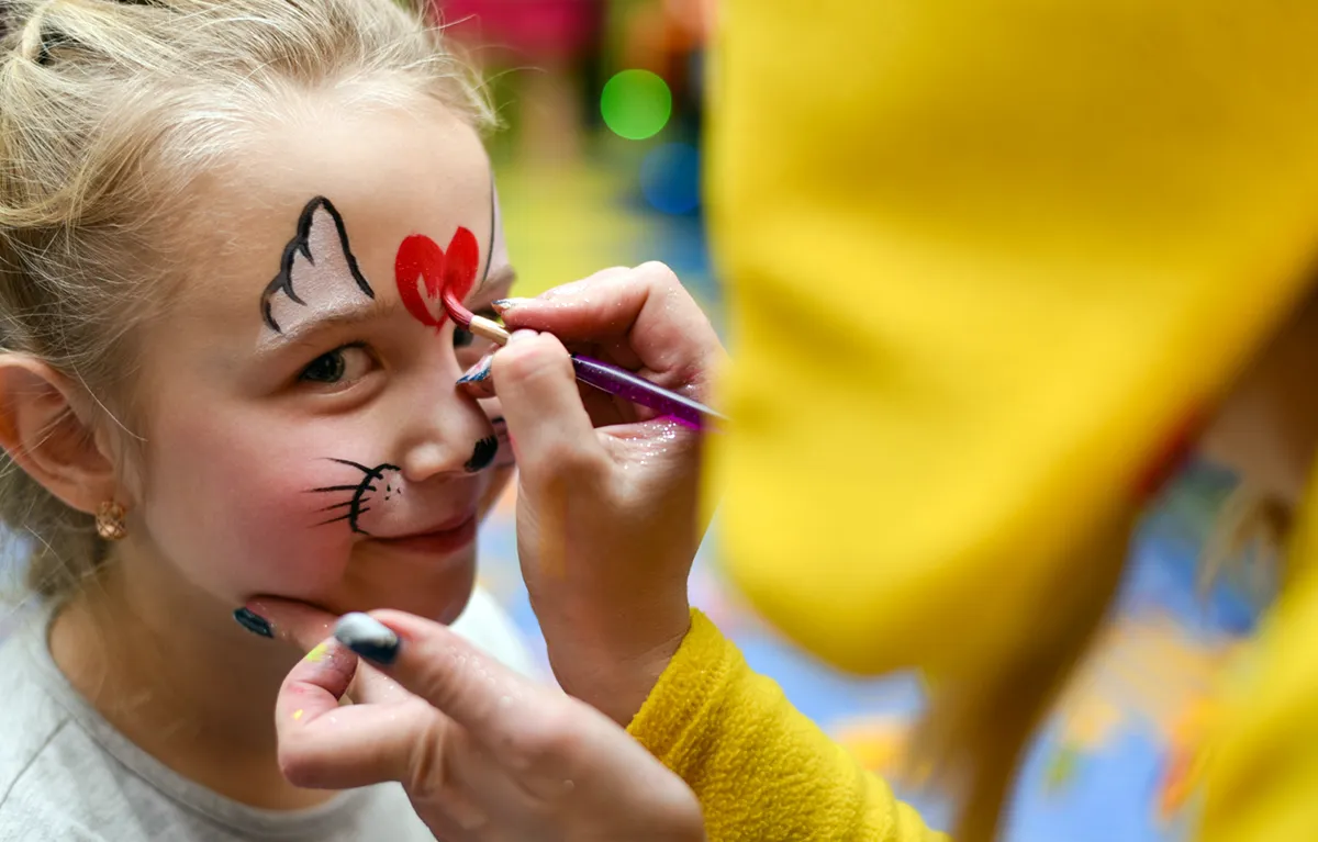 Djevojčica u krupnom planu gleda u kameru dok joj animatorica stavlja face painting; koncept djeca oboljela od malignih bolesti