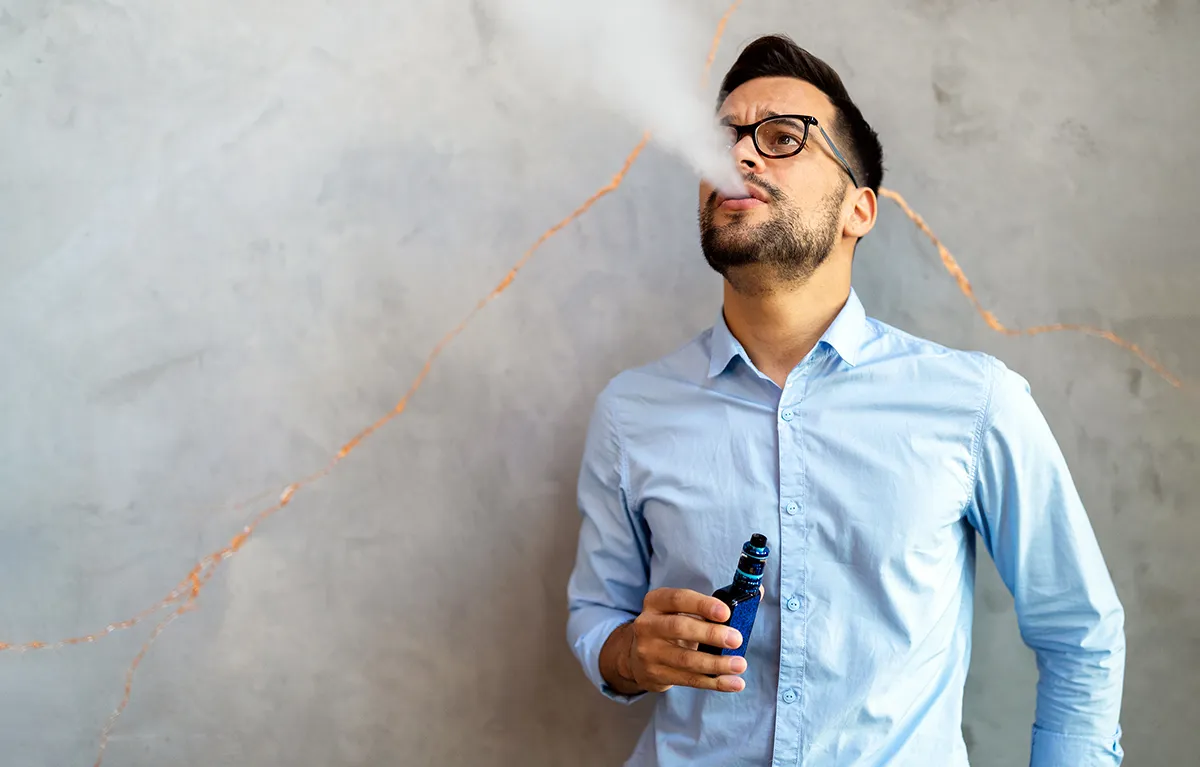 Muškarac pred sivim zidom stoji i ispuhuje dim e-cigarete; koncept što sve cigareta čini organizmu