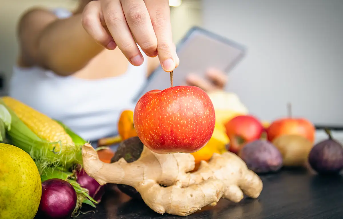 Neprepoznatljiva žena drži jabuku prema kameri, iznad stola s voćem i povrćem; prehrana kod raka dojke