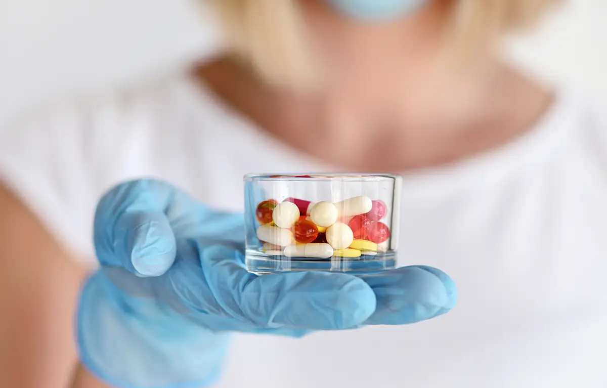 Neprepoznatljiva medicinska sestra prema kameri u ruci s medicinskom rukavicom drži staklenu posudicu s tabletama; interakcija lijekova