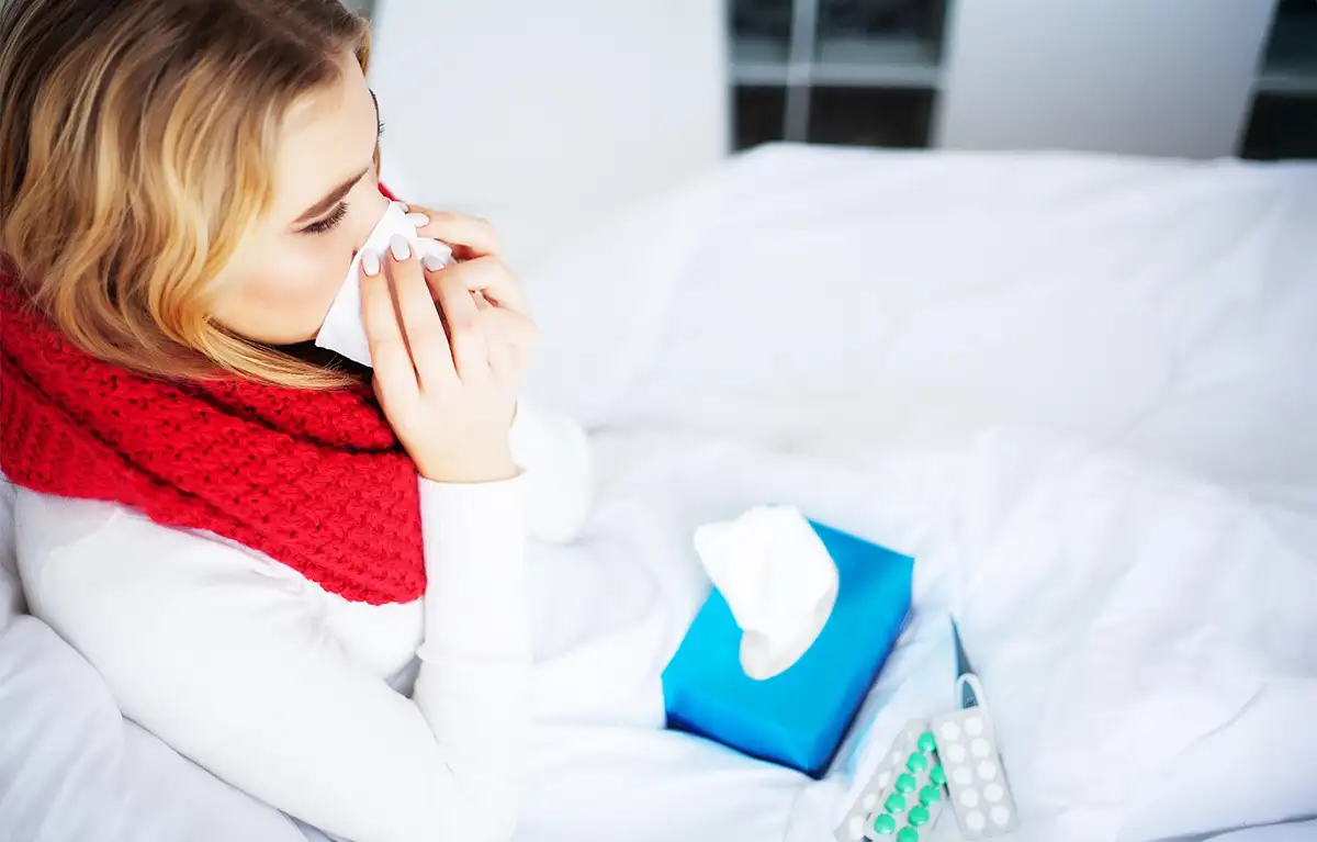 Žena u bijeloj košulji naslomnjea na krevet puše nos maramicom. Koncept: gripa i podtip svinjska gripa