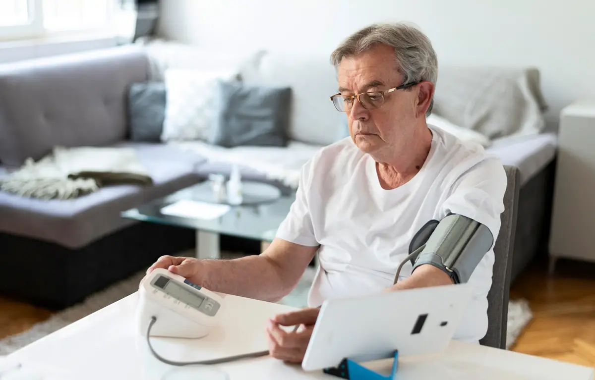 poviseni-tlak-u-Hrvatskoj-mjerenje Stariji muškarac sjedi za stolom i mjeri krvni tlak tlakomjerom; koncept povišeni tlak u Hrvatskoj.