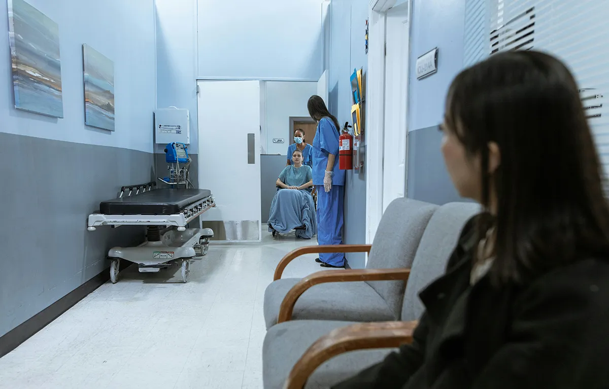 Bolnički hodnik i čekaonica, medicinske sestre voze pacijenticu u kolicima a druga pacijentica čeka i promatra; manjak liječnika u EU