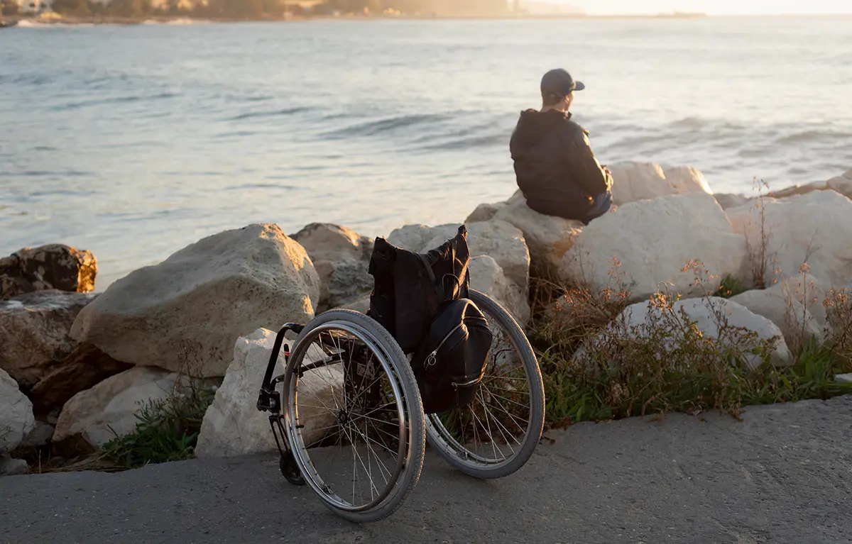 Invalidska kolica u prvom planu, muška osoba prikazana s leđa sjedi uz vodu i gleda u daljinu; Friedreichova ataksija
