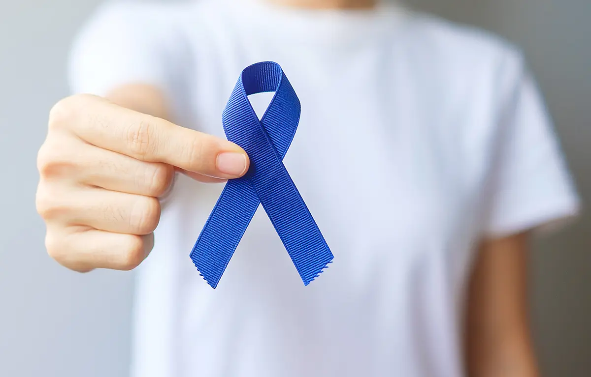 Neprepoznatljiva osoba prema kameri pokazuje tamno plavu vrpcu, simbol svjesnosti o raku debelog i završnog crijeva (rak kolona); najava za javno-zdravstvenu akciju Plavi ožujak – pregledaj se za život!, rizik za rak debelog crijeva